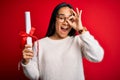 Young beautiful graduate asian woman holding university degree diploma over red background with happy face smiling doing ok sign Royalty Free Stock Photo