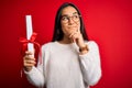 Young beautiful graduate asian woman holding university degree diploma over red background serious face thinking about question, Royalty Free Stock Photo