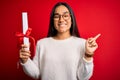 Young beautiful graduate asian woman holding university degree diploma over red background very happy pointing with hand and Royalty Free Stock Photo