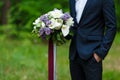 Young beautiful groom in dark blue suit standing and holding big wedding bouquet with lilac and long ribbons. Spring, park outdoor