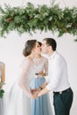 Young beautiful groom in dark trousers and white skirt and bride in white dress standing near pine arch and kissing Royalty Free Stock Photo