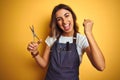 Young beautiful hairdresser woman holding scissors over yellow  background screaming proud and celebrating victory and Royalty Free Stock Photo