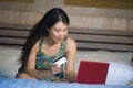 Young beautiful happy Asian American girl using credit card internet banking on laptop computer at home in bed shopping online e-c Royalty Free Stock Photo