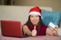 Young beautiful and happy Asian Chinese woman in Christmas Santa Claus hat holding credit card using laptop computer for internet Royalty Free Stock Photo