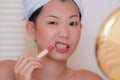 Young beautiful and happy Asian Chinese woman with towel head wrap applying makeup doing funny gesture to mirror smiling cheerful Royalty Free Stock Photo