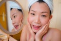 Young beautiful and happy Asian Chinese woman with towel head wrap applying makeup looking to mirror smiling cheerful preparing Royalty Free Stock Photo