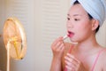 Young beautiful and happy Asian Chinese woman with towel head wrap applying makeup looking to mirror smiling cheerful preparing Royalty Free Stock Photo