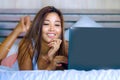 Young beautiful and happy Asian Hispanic mixed race woman smiling at home bedroom lying on bed using internet on computer laptop r Royalty Free Stock Photo