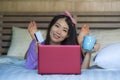 Young beautiful happy Asian Japanese woman using credit card internet banking on laptop computer at home in bed smiling shopping o Royalty Free Stock Photo