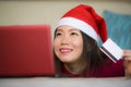 Young beautiful and happy Asian Korean woman in Christmas Santa Claus hat holding credit card using laptop computer for internet o Royalty Free Stock Photo