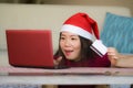 Young beautiful and happy Asian Korean woman in Christmas Santa Claus hat holding credit card using laptop computer for internet o Royalty Free Stock Photo