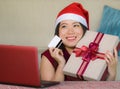 Young beautiful and happy Asian Korean woman in Santa hat holding credit card and Christmas present box using laptop computer for Royalty Free Stock Photo