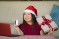 Young beautiful and happy Asian Korean woman in Santa hat holding credit card and Christmas present box using laptop computer for Royalty Free Stock Photo