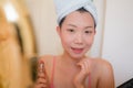 Young beautiful and happy Asian Korean woman with towel head wrap applying makeup looking to mirror smiling cheerful preparing for Royalty Free Stock Photo