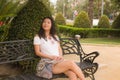 Young beautiful and happy Asian woman on park bench - lifestyle portrait of Attractive Chinese girl in Summer dress resting during Royalty Free Stock Photo