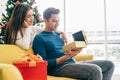 Young beautiful happy Asian woman surprises her boyfriend with a Christmas gift at home with Christmas tree in the background. Royalty Free Stock Photo
