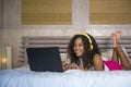 Young beautiful happy black Afro American woman at home bedroom lying cheerful on bed listening to internet music with headphones