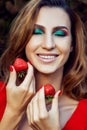 Young beautiful happy funny girl with red dress and makeup holding strawberry in summertime in the park. Royalty Free Stock Photo