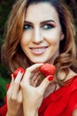 Young beautiful happy funny girl with red dress and makeup holding strawberry in summertime in the park. Royalty Free Stock Photo