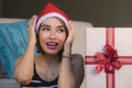 Young beautiful and happy girl in Santa hat holding Christmas present box with ribbon smiling cheerful and excited at home couch r Royalty Free Stock Photo