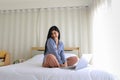 Young beautiful happy long black hair asian female wearing blue long sleeve shirt smiling and sitting on white sheet bed using Royalty Free Stock Photo