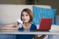 Young beautiful and happy woman at home living room holding credit card using laptop computer for banking and online shopping smil Royalty Free Stock Photo