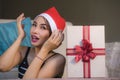 Beautiful and happy woman in Santa hat holding Christmas present box with red ribbon smiling excited at home living room receiving Royalty Free Stock Photo