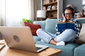 Young beautiful hipster woman sitting on sofa at home listening music on wireless headset and reading a book. Freelance female Royalty Free Stock Photo