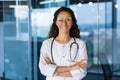 Young beautiful hispanic female doctor with arms crossed smiling and looking at camera, female doctor in medical gown Royalty Free Stock Photo