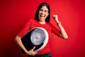 Young beautiful hispanic fitness woman holding scale for healthy weight over red background screaming proud and celebrating Royalty Free Stock Photo