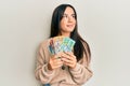 Young beautiful hispanic girl holding australian dollars smiling looking to the side and staring away thinking