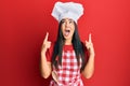 Young beautiful hispanic girl wearing baker uniform and cook hat amazed and surprised looking up and pointing with fingers and Royalty Free Stock Photo