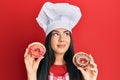 Young beautiful hispanic girl wearing chef hat holding homemade doughnut smiling looking to the side and staring away thinking Royalty Free Stock Photo