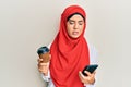 Young beautiful hispanic girl wearing islamic hijab drinking a coffee usign smartphone in shock face, looking skeptical and