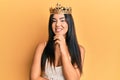 Young beautiful hispanic girl wearing queen crown smiling looking confident at the camera with crossed arms and hand on chin Royalty Free Stock Photo
