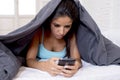 Young beautiful hispanic mobile phone addict woman in bed with blanket over head texting internet surfing