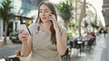 Young beautiful hispanic woman assistant talking on smartphone reading notebook at coffee shop terrace Royalty Free Stock Photo