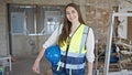 Young beautiful hispanic woman builder smiling confident holding hardhat at construction site Royalty Free Stock Photo