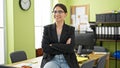 Young beautiful hispanic woman business worker sitting on table with arms crossed gesture smiling at office Royalty Free Stock Photo