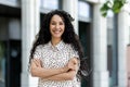 Young beautiful hispanic woman with curly hair smiling and looking at camera, businesswoman with arms crossed outside Royalty Free Stock Photo