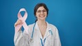 Young beautiful hispanic woman doctor holding breast cancer awareness pink ribbon over isolated blue background Royalty Free Stock Photo