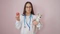 Young beautiful hispanic woman doctor smiling confident holding teddy bear and apple over isolated pink background Royalty Free Stock Photo