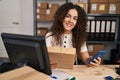 Young beautiful hispanic woman ecommerce business worker using smartphone holding package at office Royalty Free Stock Photo