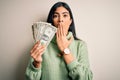 Young beautiful hispanic woman holding a pack of one dollar bunch of dollars cover mouth with hand shocked with shame for mistake, Royalty Free Stock Photo