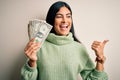 Young beautiful hispanic woman holding a pack of one dollar bunch of dollars pointing and showing with thumb up to the side with Royalty Free Stock Photo