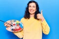 Young beautiful hispanic woman holding paintbrush and palette smiling happy and positive, thumb up doing excellent and approval Royalty Free Stock Photo