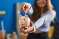 Young beautiful hispanic woman inserting coin on piggy bank standing at new home Royalty Free Stock Photo