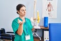 Young beautiful hispanic woman physiotherapist holding anatomical model of knee at rehab clinic