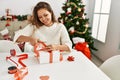 Young beautiful hispanic woman preparing christmas gift sitting on table at home Royalty Free Stock Photo