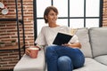 Young beautiful hispanic woman reading book and drinking coffee sitting on sofa at home Royalty Free Stock Photo